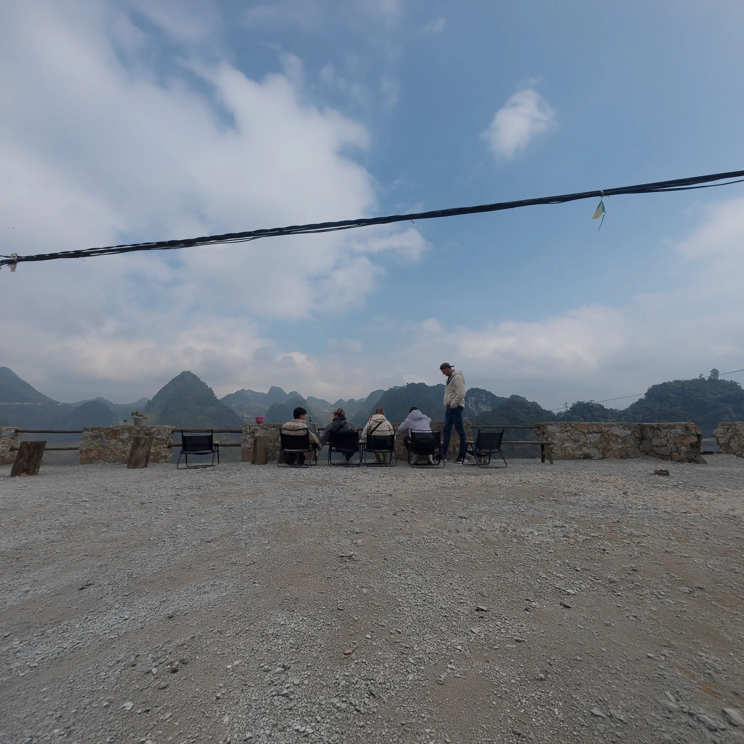 Travellers sit in chairs at a gravel viewpoint terrace overlooking the dramatic karst limestone peaks of the Ha Giang Loop, Vietnam.