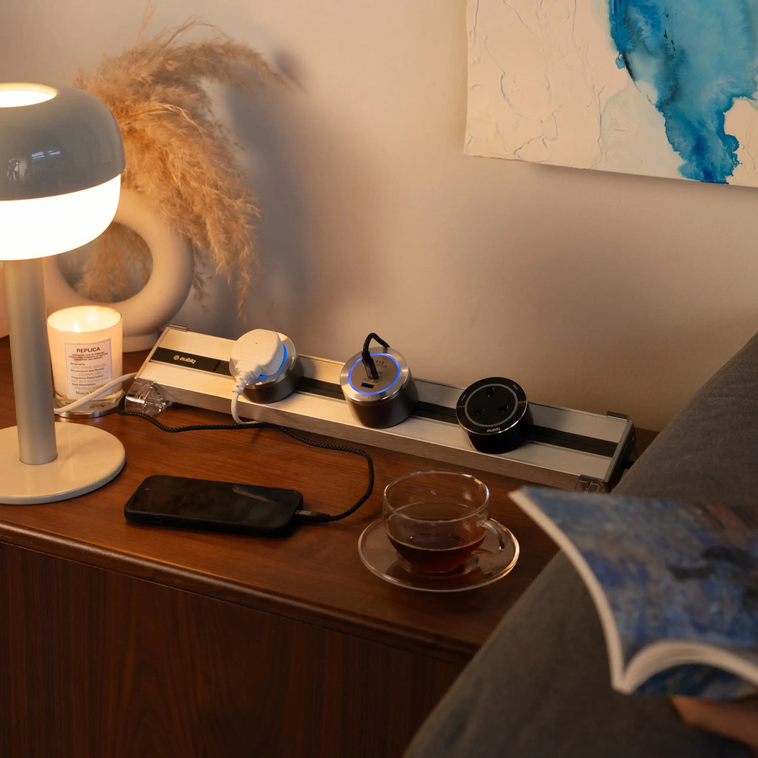 A Eubiq bedside power track discreetly charges a phone and powers a lamp for reading in a serene setting.