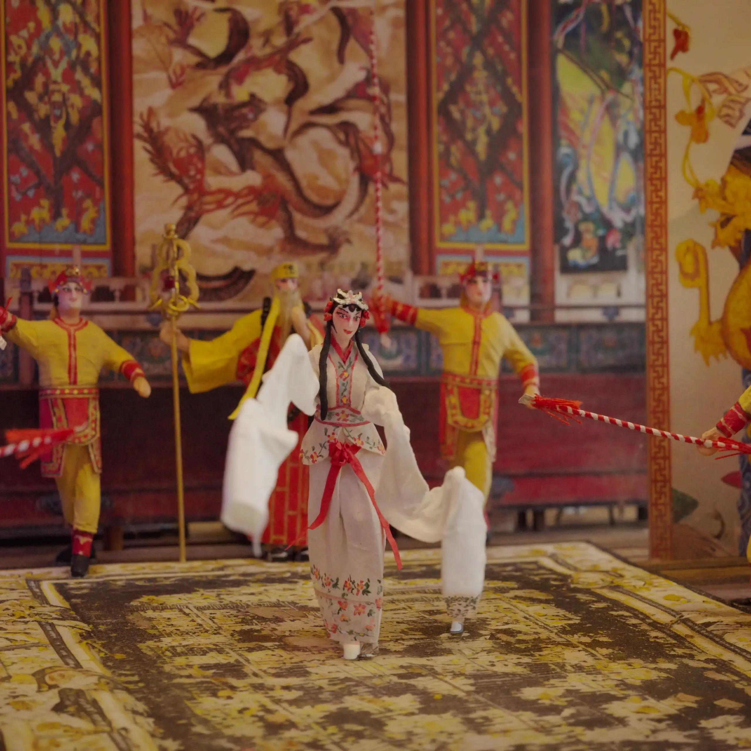 Wilfred Cheah smiles warmly in front of an intricate miniature diorama depicting a Chinese opera stage performance with tiny costumed figures and an audience below.
