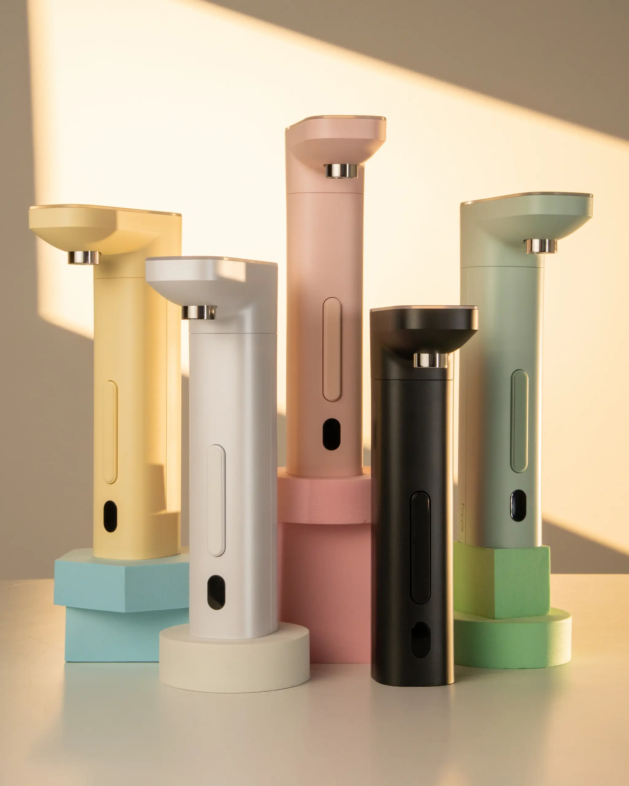 Happie dispensers in five colors arranged in blocks, shown in warm sunset lighting to highlight the variety of aesthetic options available.